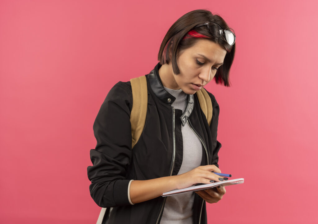 concentrated young student girl wearing glasses on head and back bag holding pen and note pad looking at note pad isolated on pink background with copy space
