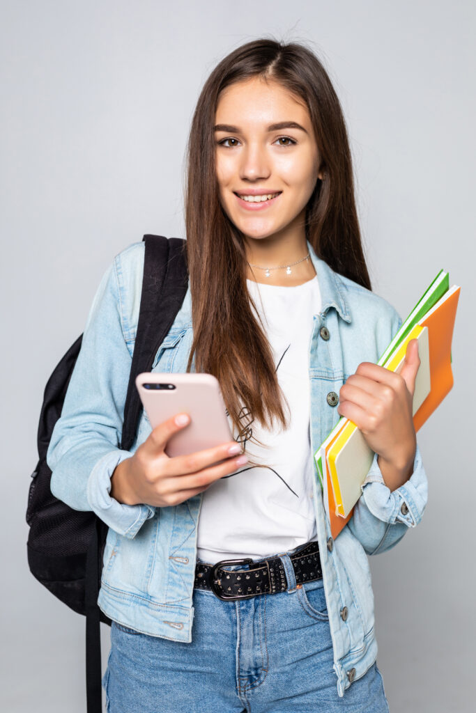 Portrait of happy young woman standing with backpack holding books and mobile phone isolated on white background